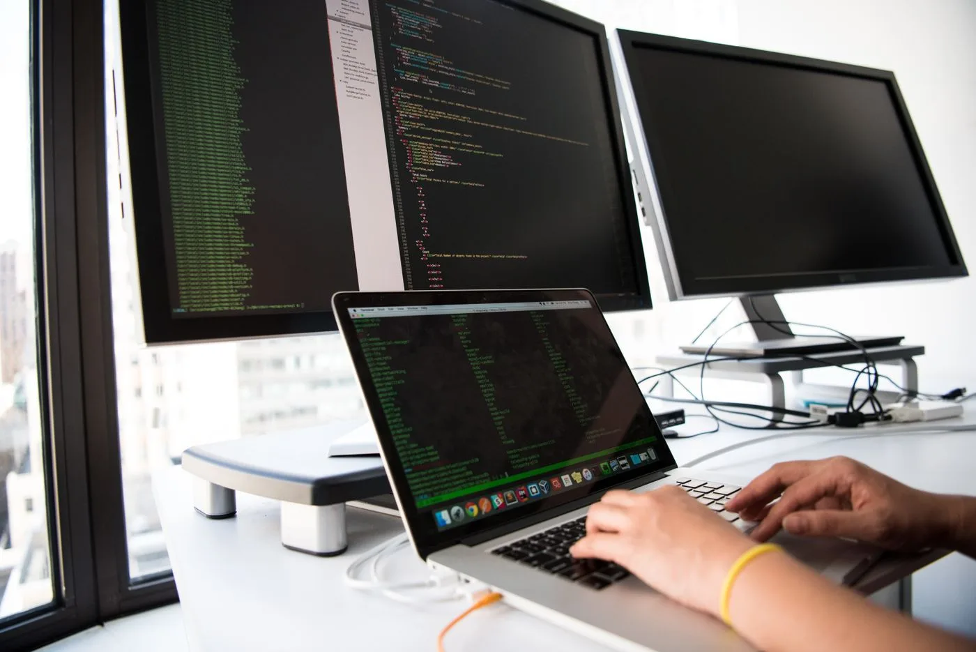 A developer writes code on a laptop in front of multiple monitors in an office setting