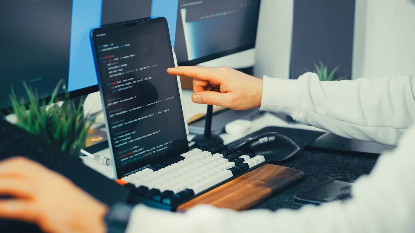 A programmer working on code with a laptop and monitor setup in an office