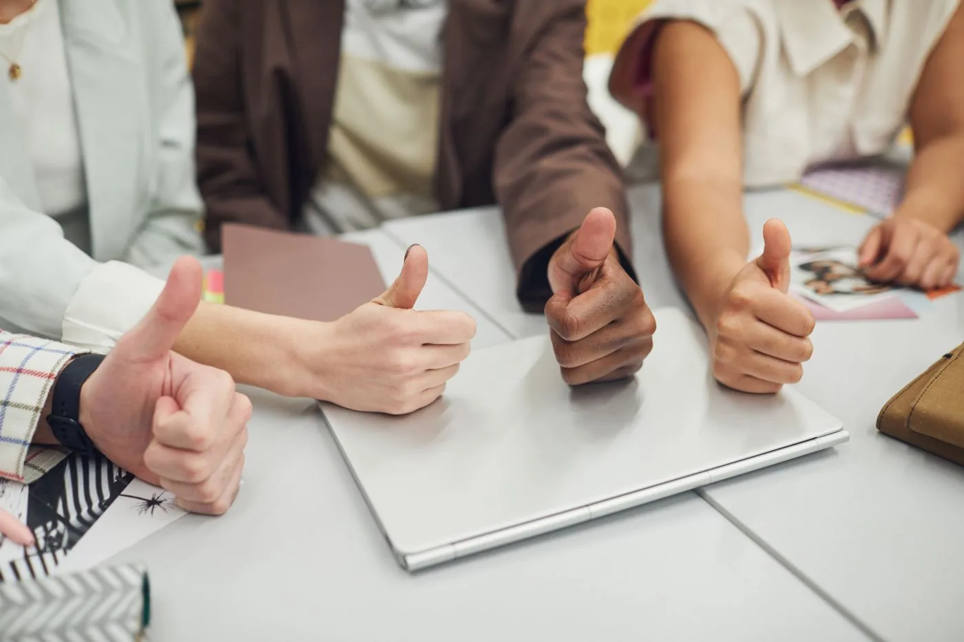 Four diverse hands showing thumbs up over a laptop on a desk