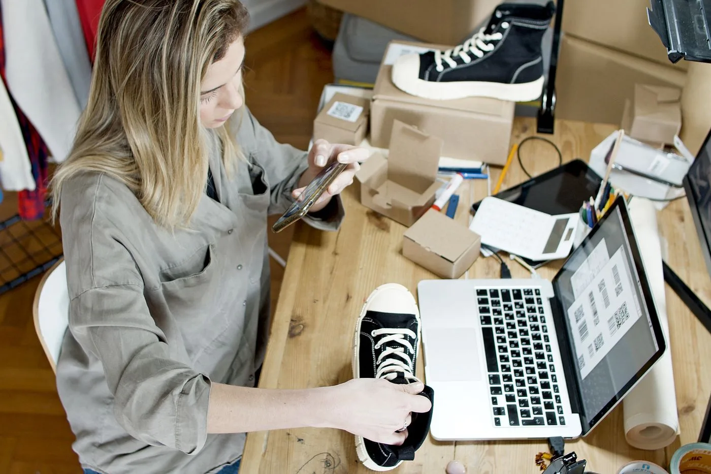 Woman photographing shoes for online sale in home workspace with laptop and packages