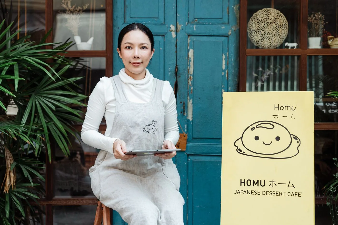 A woman holding a tablet sits outside a Japanese dessert cafe with a welcoming smile