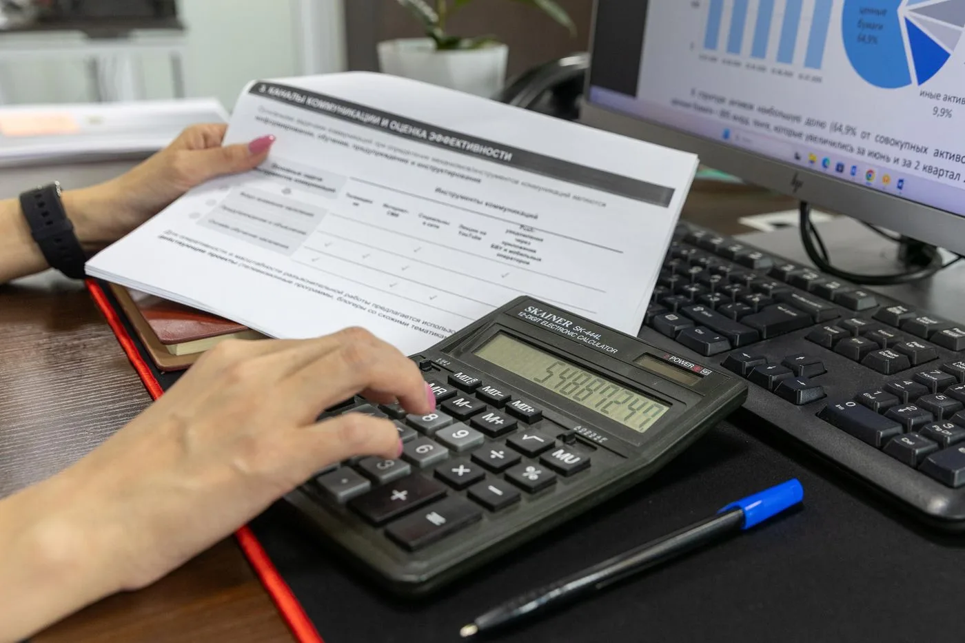 A person calculates financial data using a calculator and document, working at an office desk