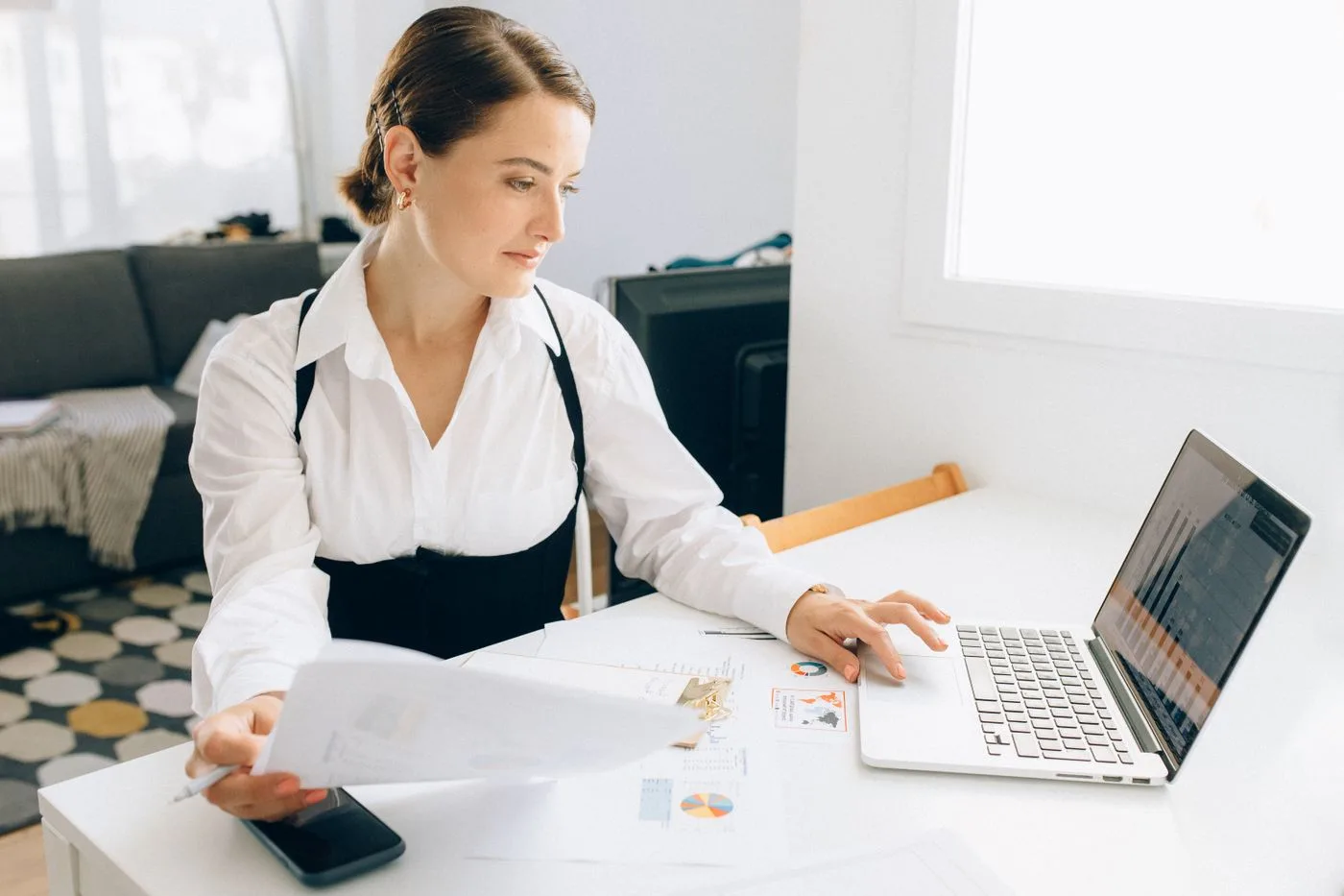 Woman at desk reviewing business reports on laptop, holding papers in a modern office setting