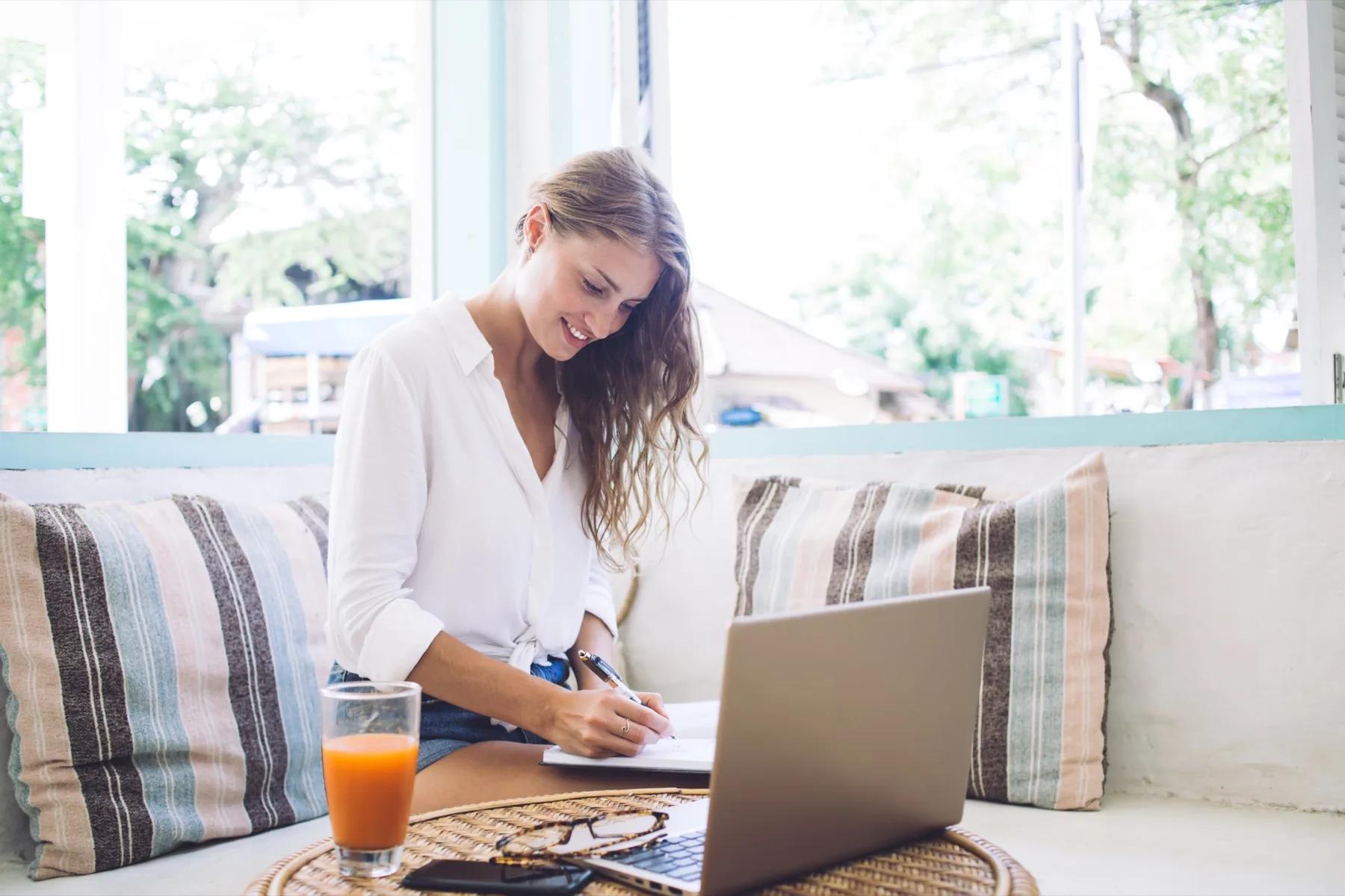 Woman writing notes by a sunny window with her laptop open, drafting copy