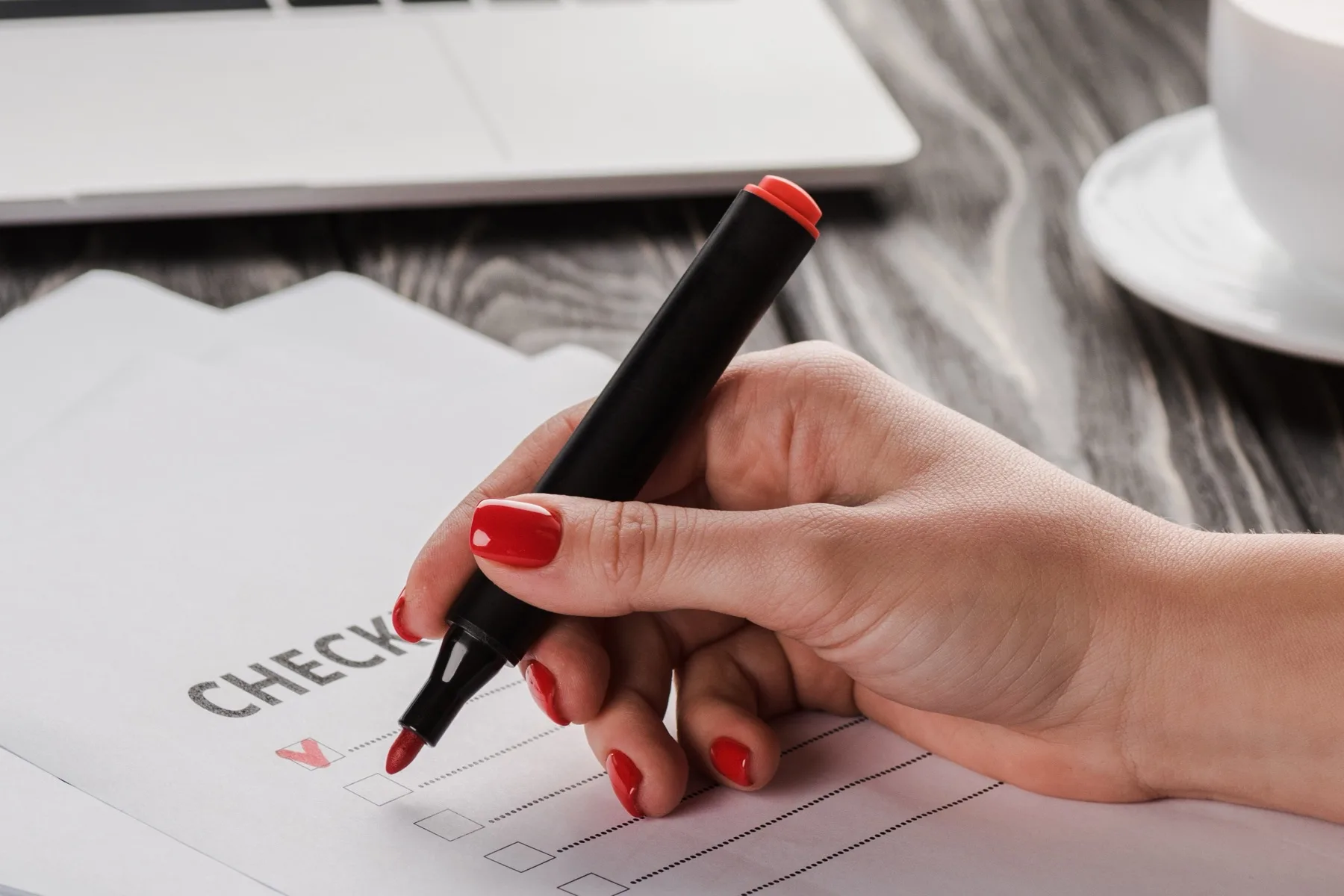 Hand marking up a printed checklist with a red marker beside a laptop and coffee cup