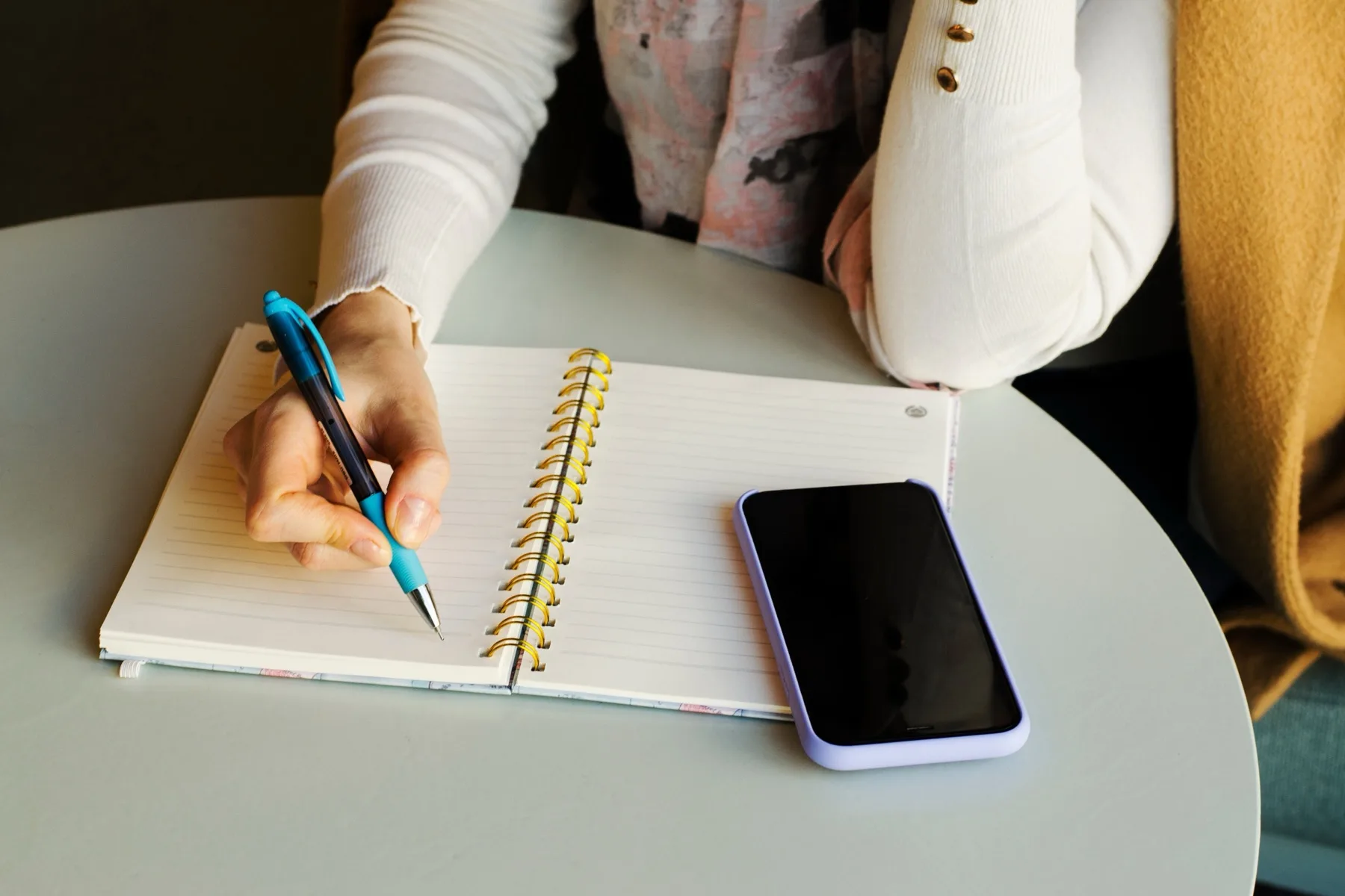Woman writing a caption draft by hand in a spiral notebook, with her phone face down beside it
