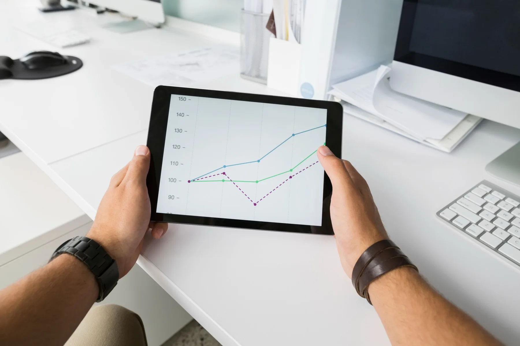 Hands holding a tablet showing line charts at a white office desk