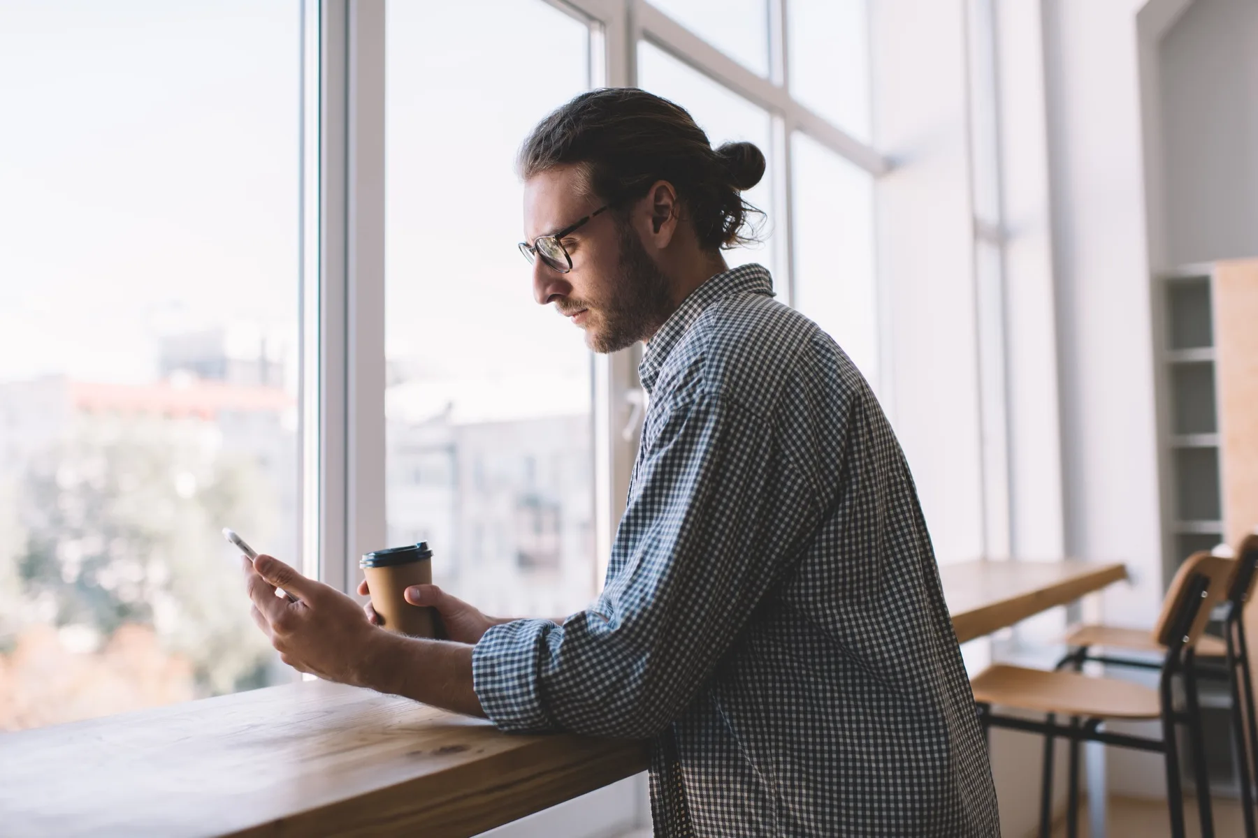 Man with a coffee reading his phone at a window counter, deciding whether to keep reading