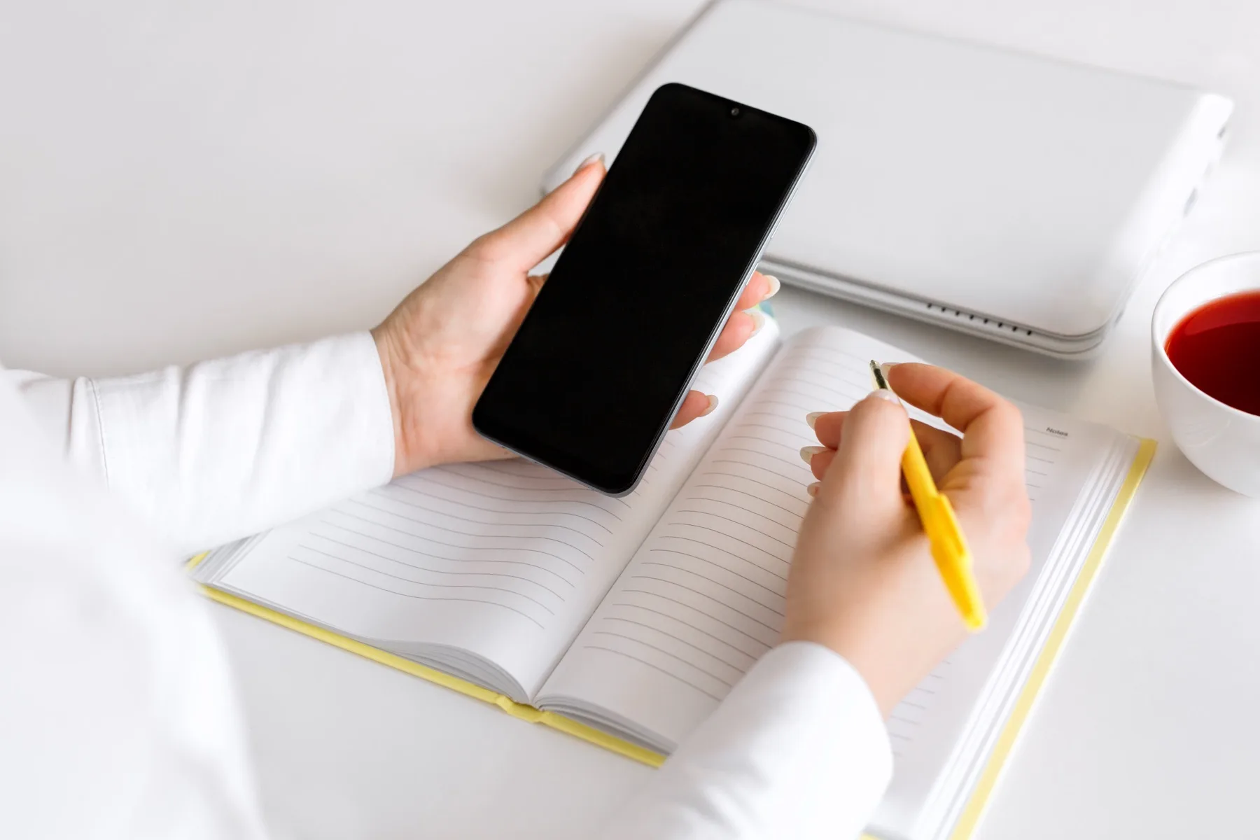 Woman holding a smartphone while noting ideas in an open notebook at a white desk