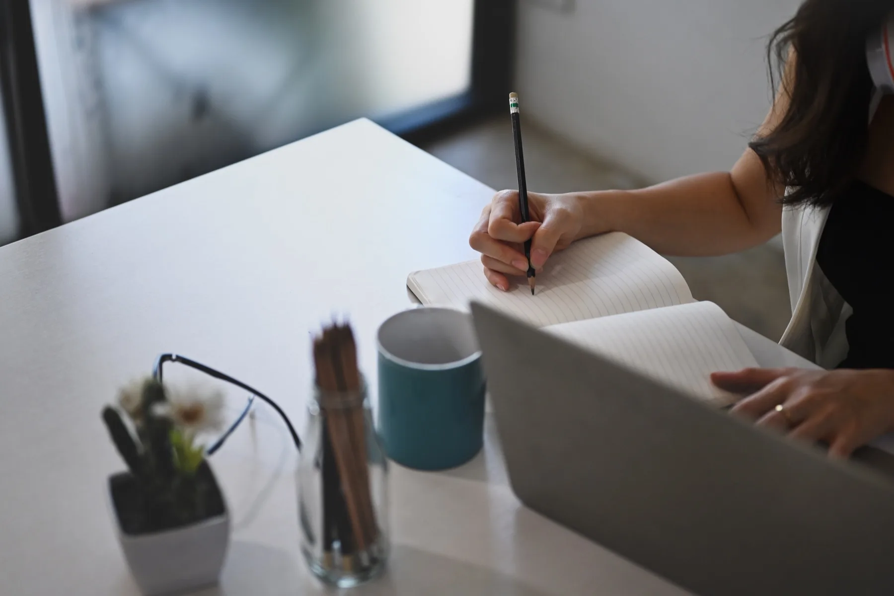 Woman writing in a notebook at a white desk next to an open laptop, a mug and a pen pot
