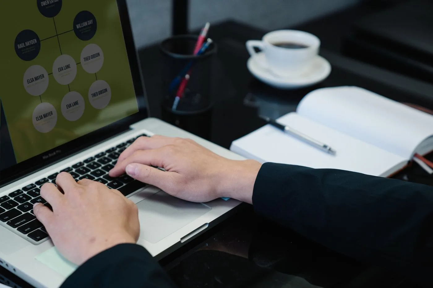 Close-up of hands typing on a laptop displaying an organizational chart, a notebook, and coffee nearby