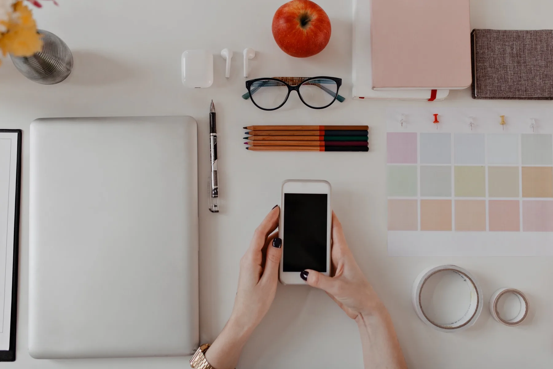 Overhead view of a tidy desk with a laptop, notebook, colour swatches and hands holding a smartphone, ready to write an Instagram caption