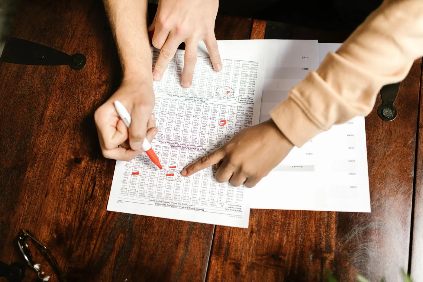 Two hands reviewing and marking financial documents with a red pen on a wooden table, top view