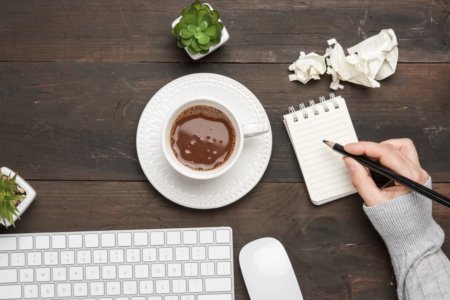 Writer drafting in a notepad next to a keyboard and coffee on a wooden desk