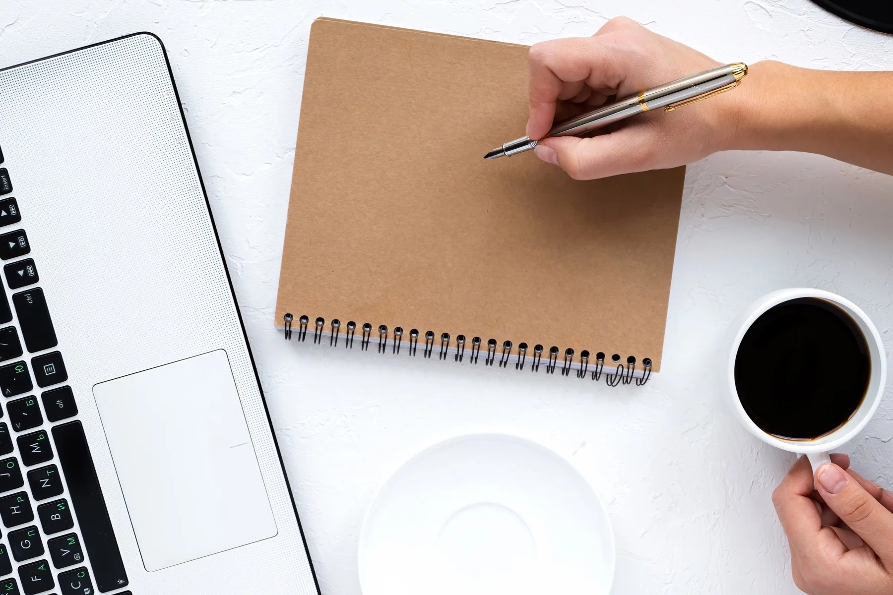 Hand writing in a kraft notebook next to a laptop and coffee