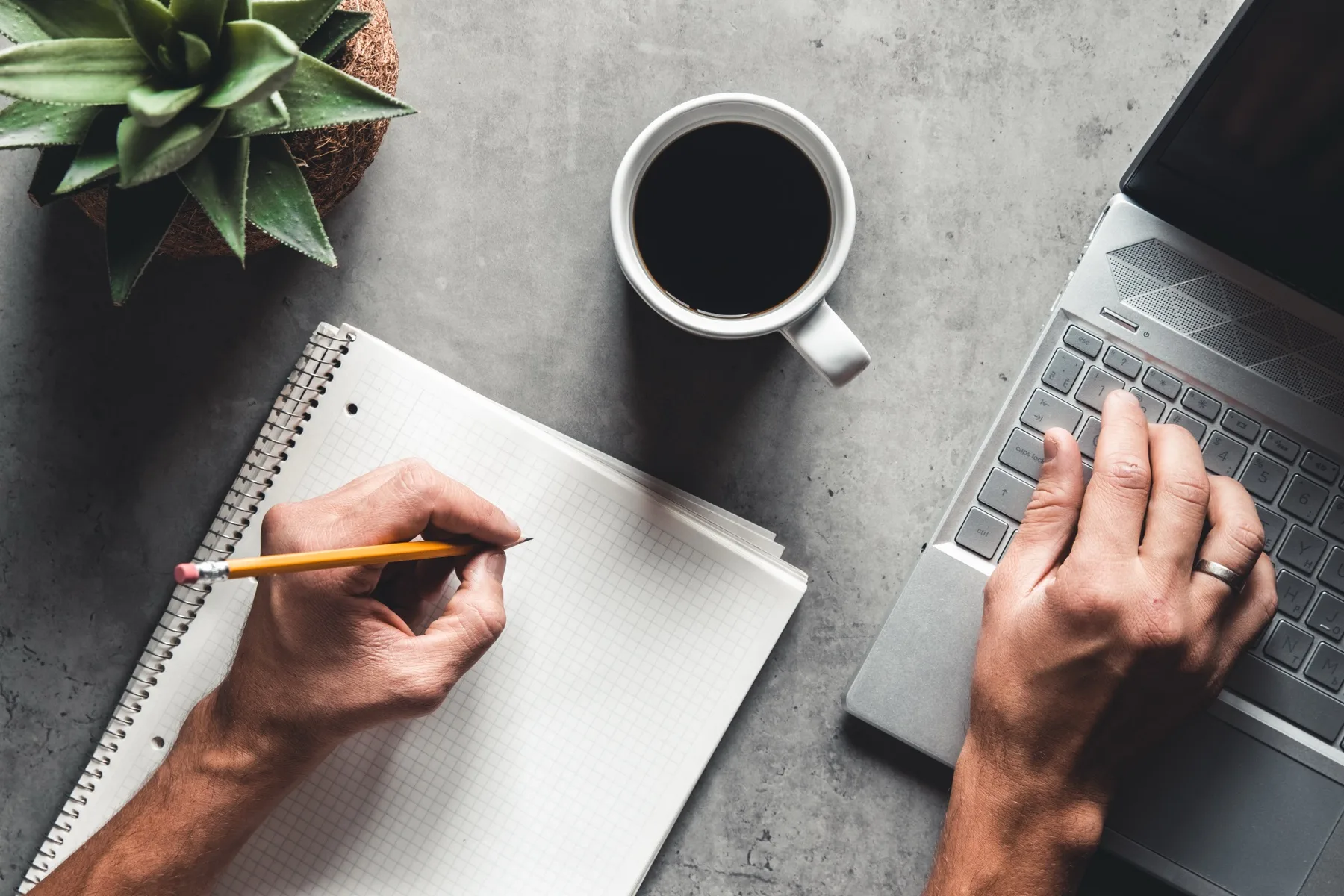 Person writing in a notebook beside a laptop and coffee on a grey desk