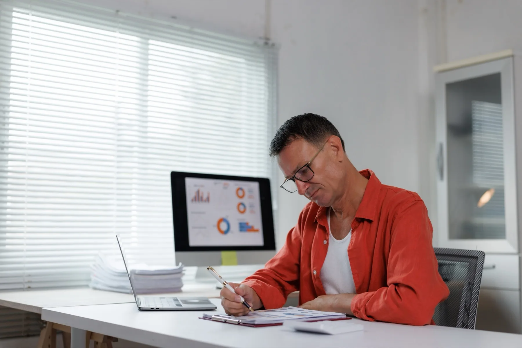 Man writing up figures at a desk in front of a monitor showing charts