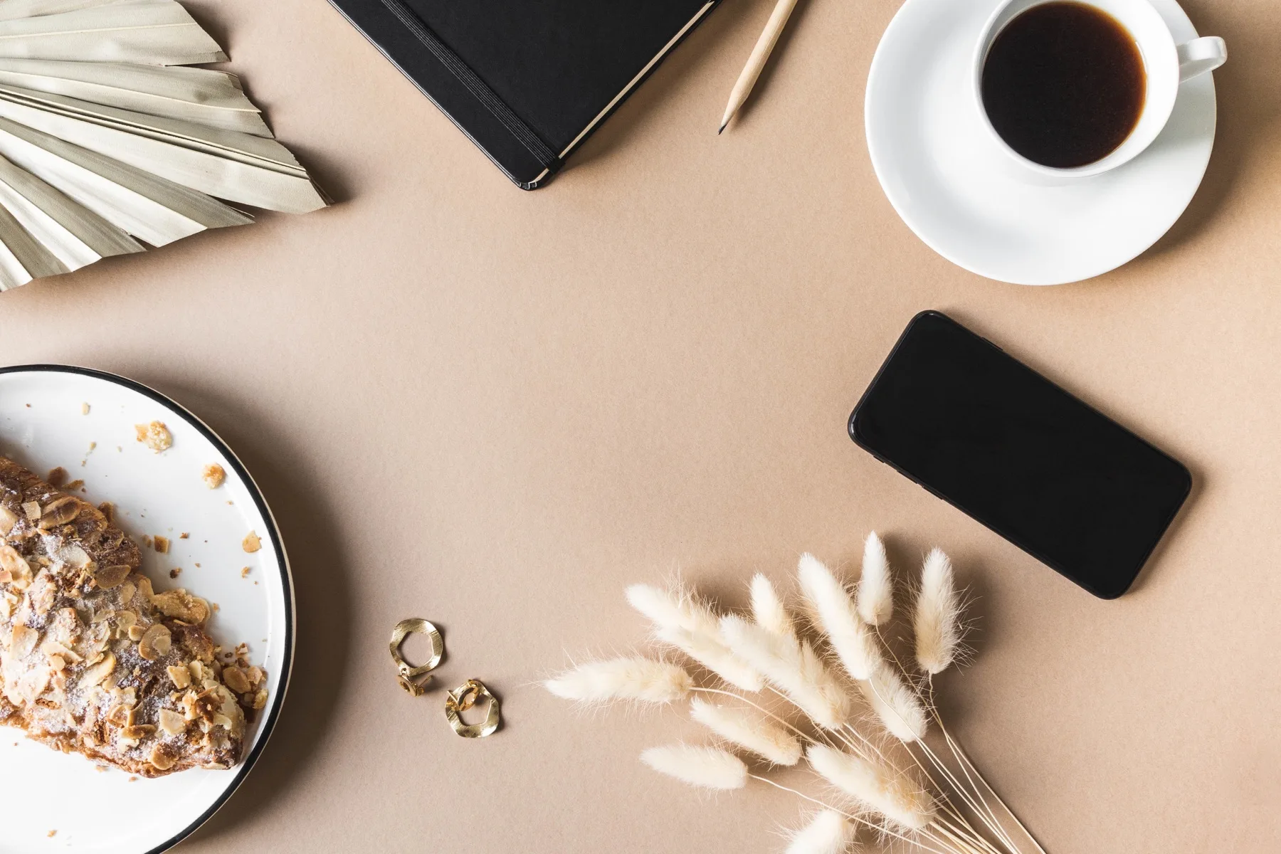 Flat lay of a phone, coffee, pastry and dried flowers on a beige table
