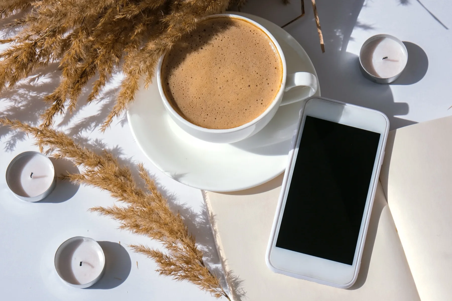 Smartphone next to a coffee, candles and dried grass on a white surface