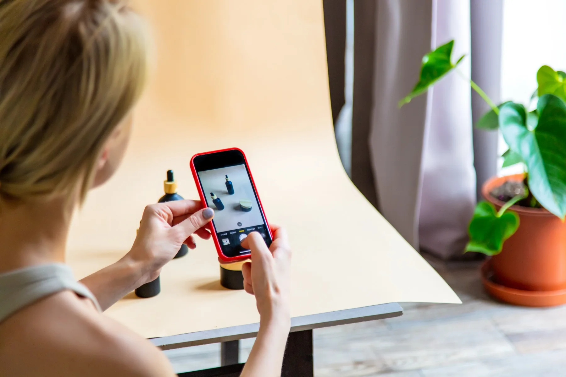 Woman photographing skincare bottles on her phone at a desk