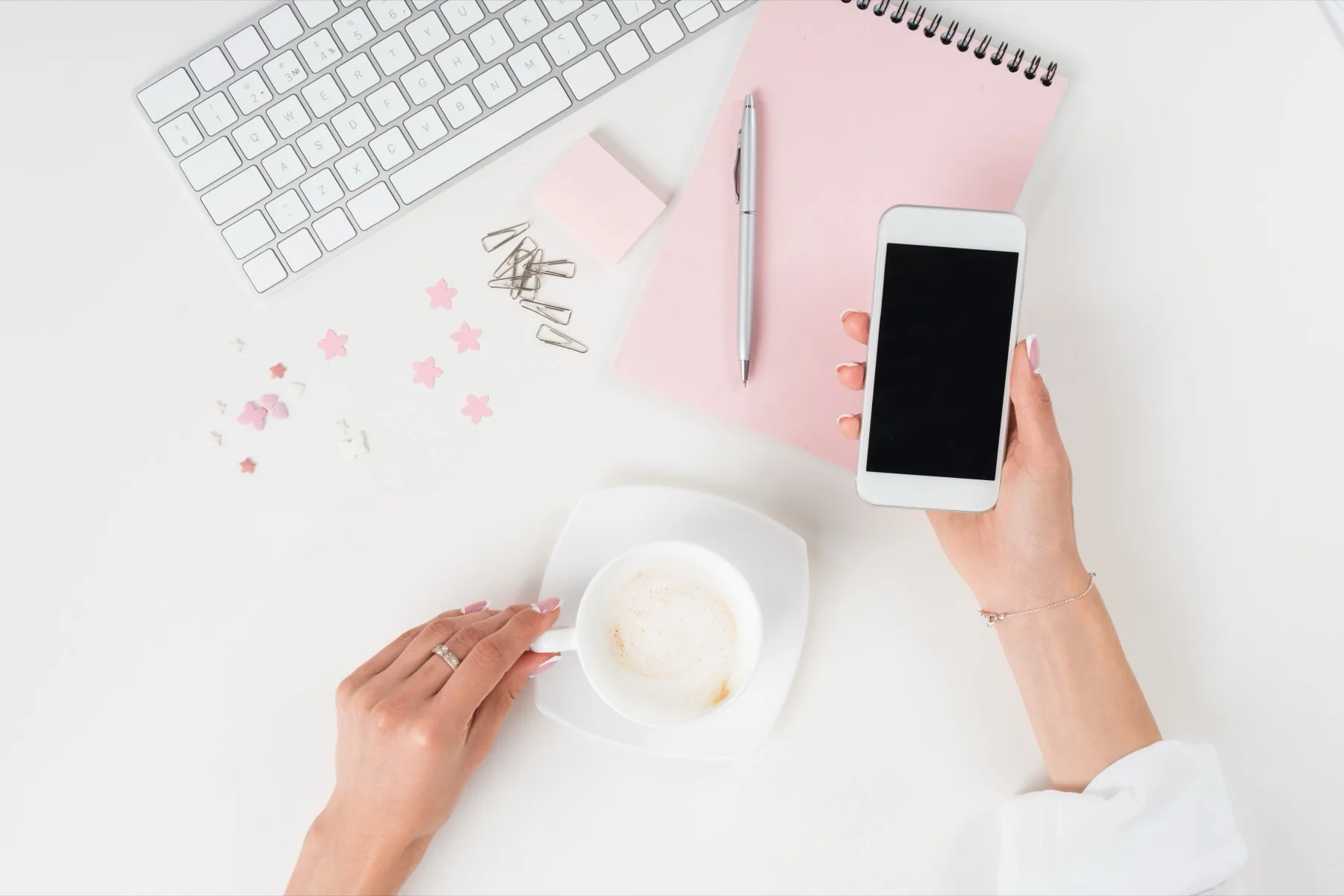 Overhead view of a white desk with a keyboard, a pink notebook and coffee, and hands holding a smartphone ready to write a caption