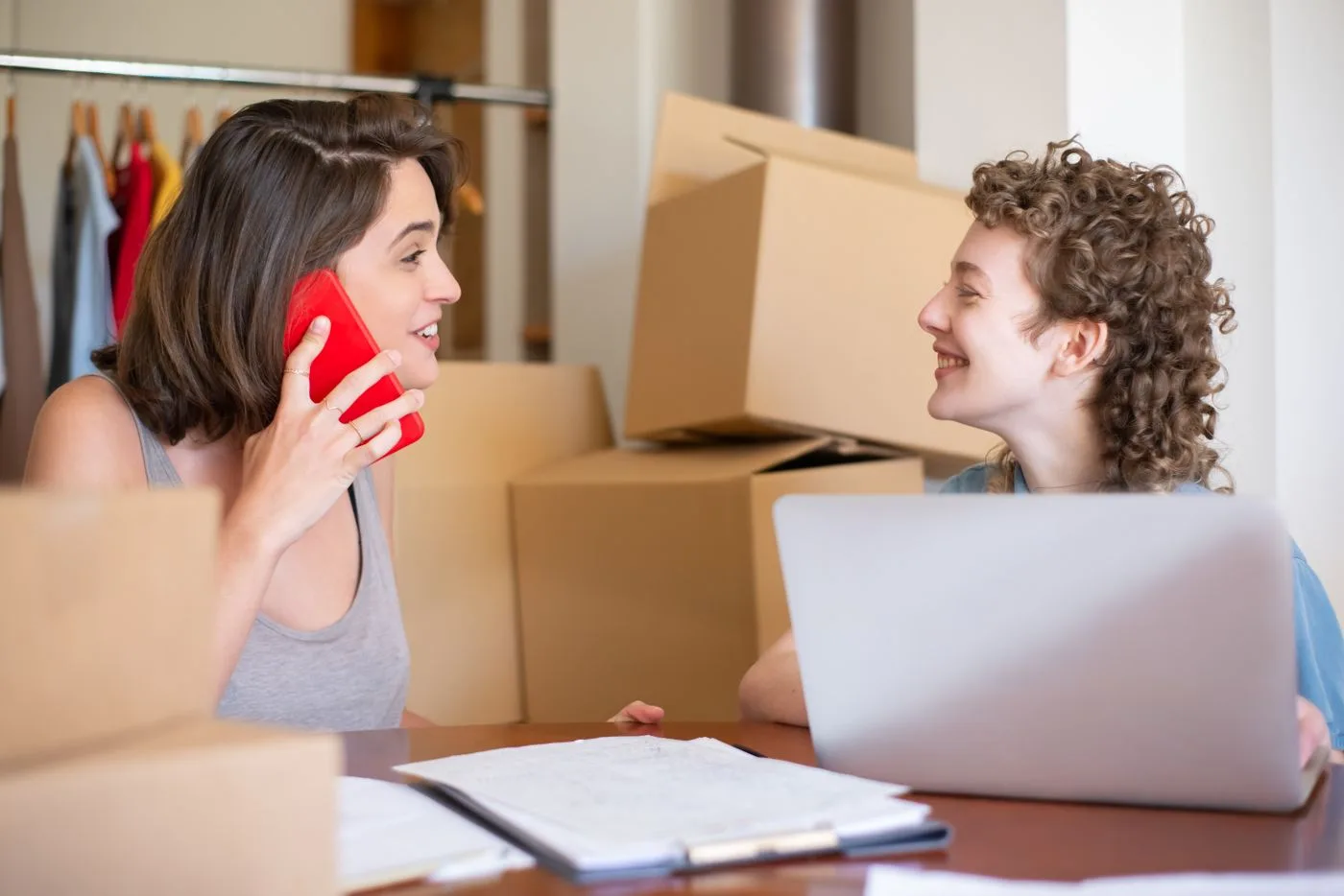 Two women managing their online clothing store using a laptop and phone, surrounded by boxes